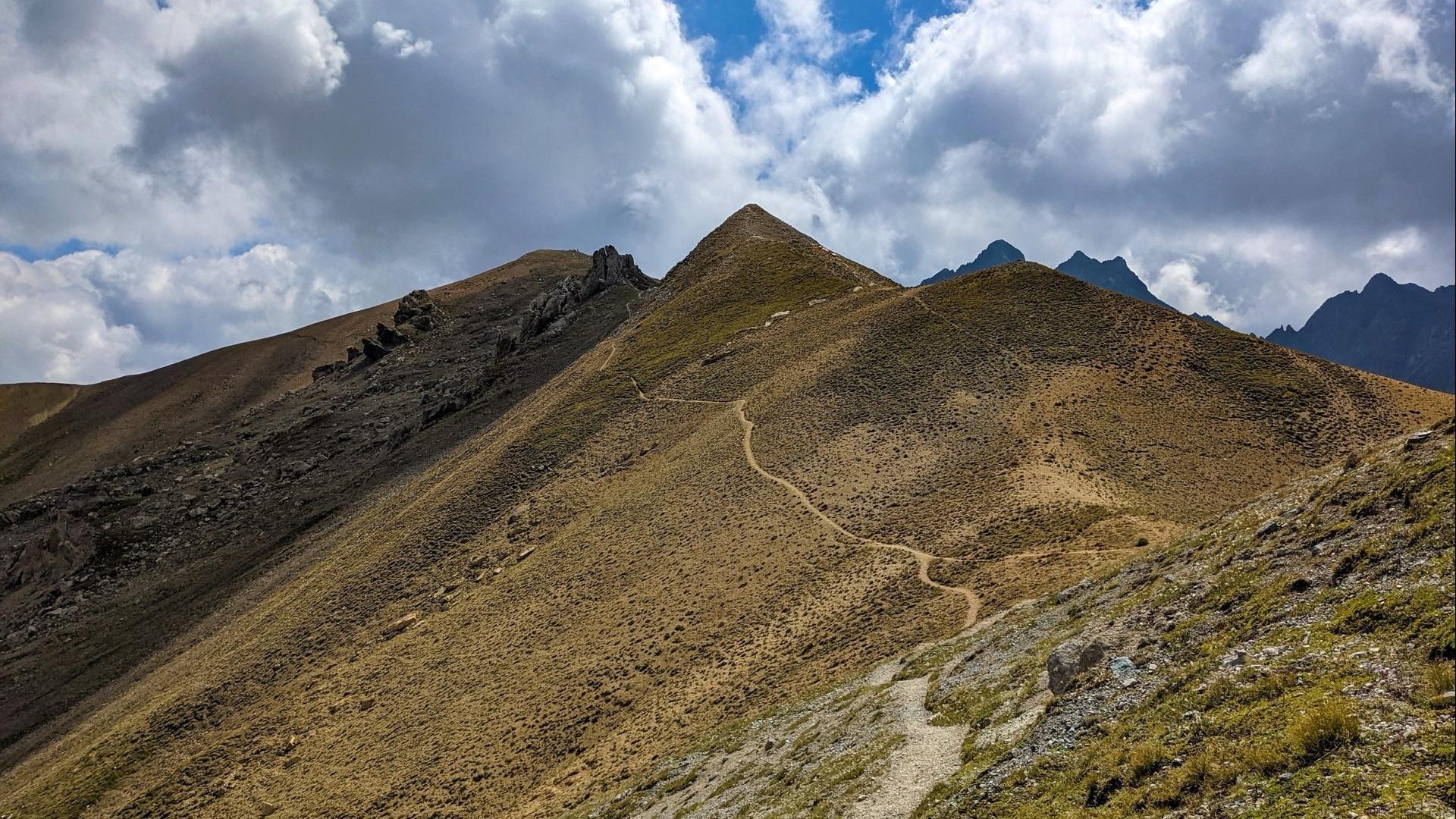 A path along a mountain ridge climbing toward a summit under a clouded sky.