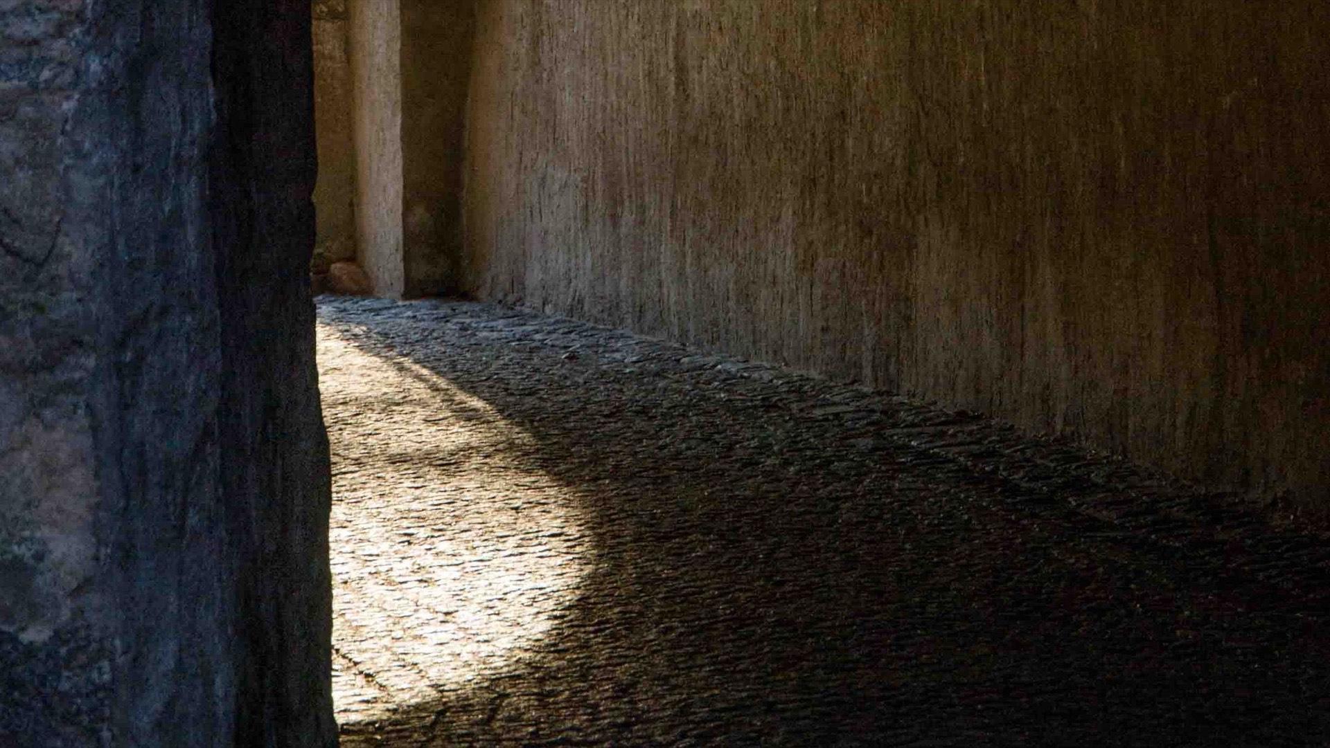A dark stone passage, with sunlight pooling on the cobbles beyond the arch.
