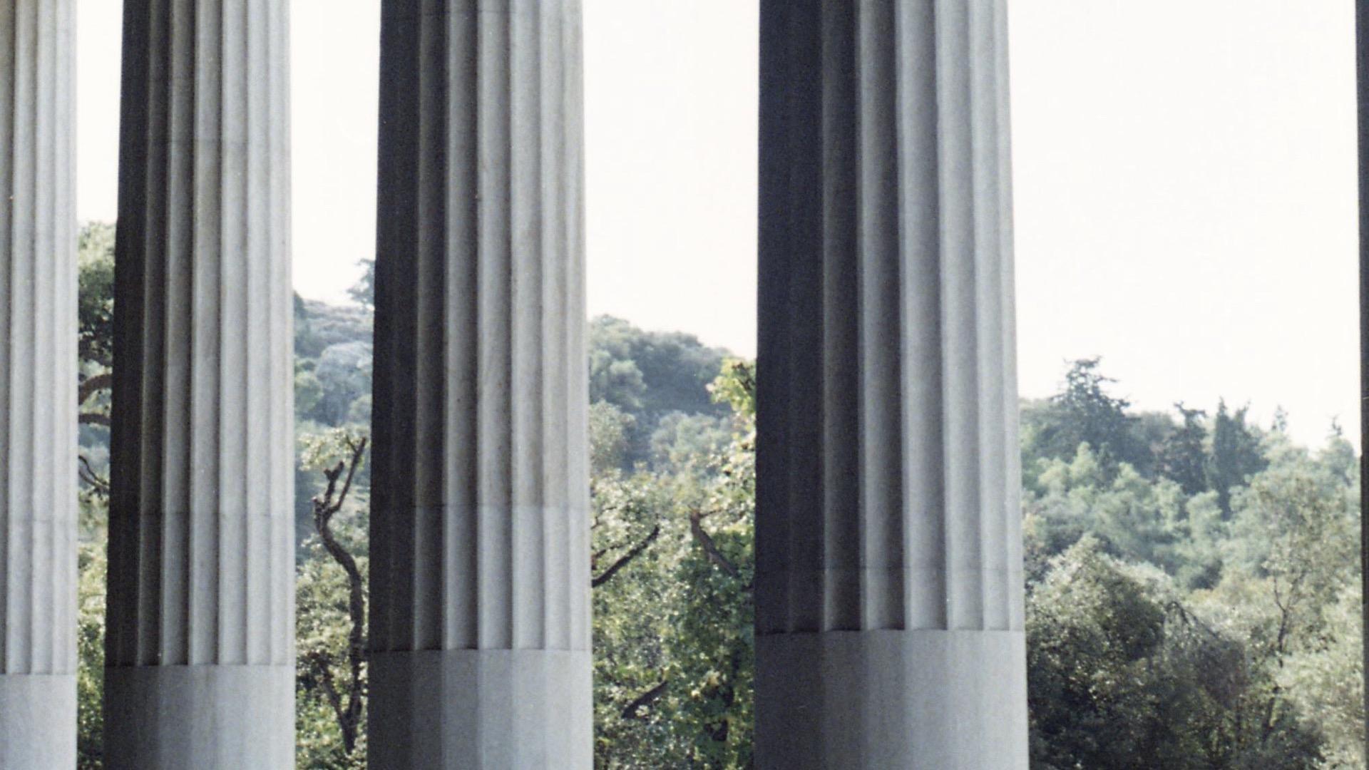 A row of fluted stone columns along the edge of a terrace, trees beyond.
